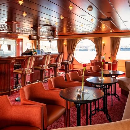 Elegante Salon-Bar auf der MS Switzerland mit orangen und beigen Sitzgelegenheiten, dunkler Holzverkleidung, runden Bullaugen und einer Theke.