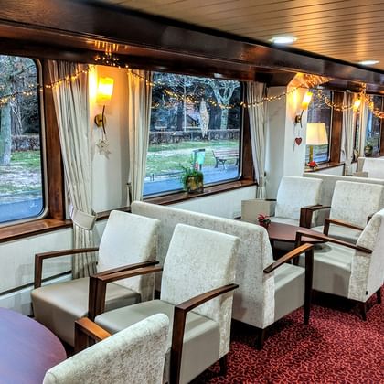 Innenraum des MS Classica Salons mit Reihen weißer Sessel, rotem Teppich, großen Fenstern mit Vorhängen und warmer Beleuchtung. Bäume draußen sichtbar.