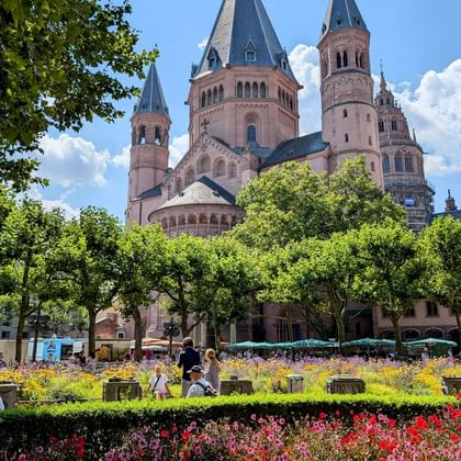 Mainz Cathedral with six red sandstone towers rising above a colorful flower garden. Visitors stroll through the landscaped area with trees.