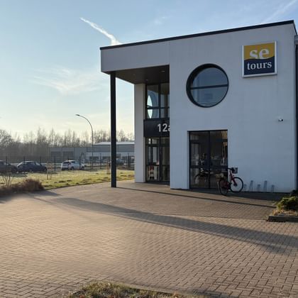 Modern white office building with SE Tours yellow sign, circular window, and covered entrance. Bicycles parked outside, paved parking area visible.