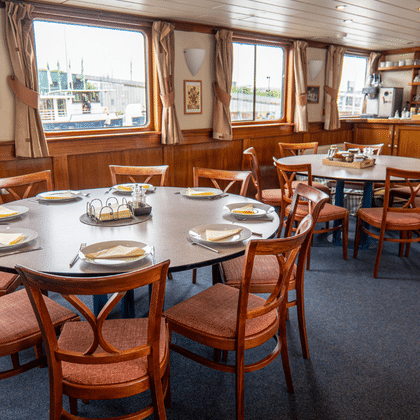 Speisesaal auf dem Schiff Clair de Soleil mit runden und rechteckigen Tischen, Holzstühlen, Panoramafenstern und Barbereich.