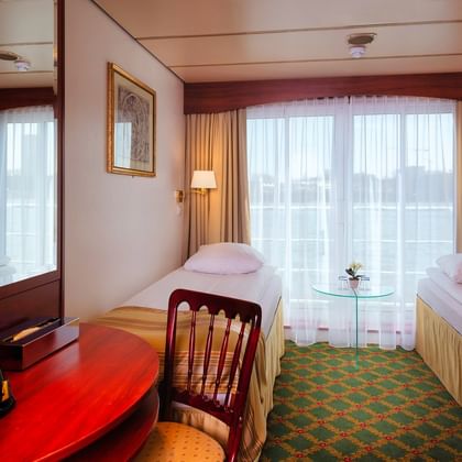 Twin cabin on MS Switzerland with two single beds, large windows with curtains, green patterned carpet, and wooden furniture including desk and chair.