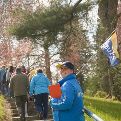 Reiseleiter in blauer Jacke mit Flagge und Mappe führt Touristengruppe einen von Bäumen gesäumten Weg mit grünem Grashang hinauf.