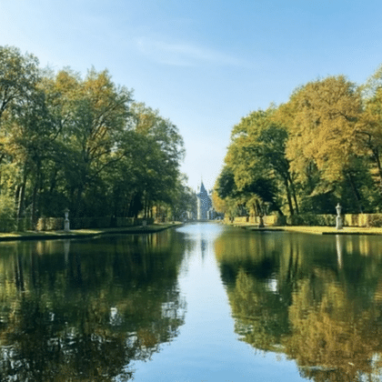 Tranquil park pond reflecting autumn trees in golden and green hues. A church spire is visible in the distance under a blue sky.