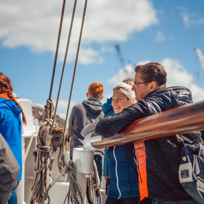 Passengers on the deck of sailing ship Mare Frisium enjoying a sunny day. People in casual jackets lean against wooden railings with rigging visible.