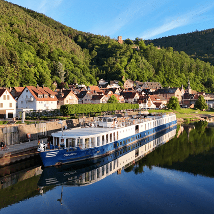 Blau-weißes Flusskreuzfahrtschiff Casanova am Kai eines Dorfes mit traditionellen Häusern, grünen bewaldeten Hügeln und ruhigen Wasserspiegelungen.
