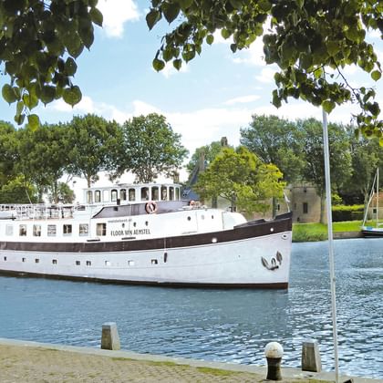 Weißes Passagierschiff Floor van Aemstel mit dunklem Rumpf auf Wasserstraße, eingerahmt von Baumzweigen. Historische Gebäude im Hintergrund.