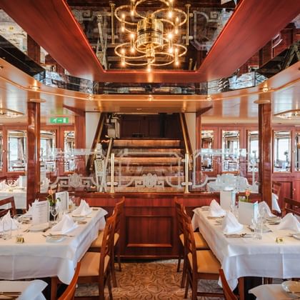 Elegantes Restaurant an Bord der MS Switzerland mit weiß gedeckten Tischen, hölzernem Schaufelrad und Messingkronleuchter unter Spiegeldecke.