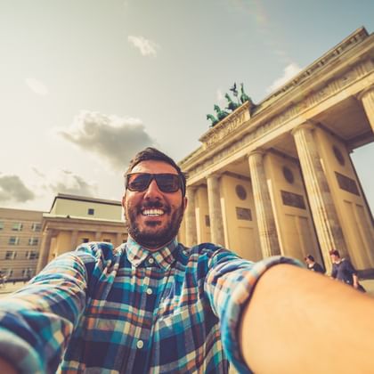 Lächelnder Mann mit Sonnenbrille macht Selfie vor dem Brandenburger Tor in Berlin. Das neoklassizistische Monument mit Säulen ist hinter ihm sichtbar.