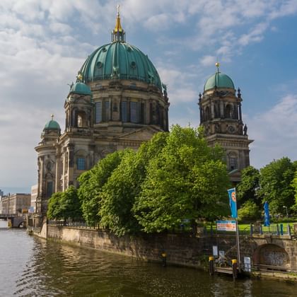 Berliner Dom mit charakteristischen grünen Kupferkuppeln an der Spree, umgeben von üppigen grünen Bäumen unter bewölktem Himmel.