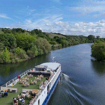 Luftaufnahme des Flusskreuzfahrtschiffs Casanova mit Sonnendeck und Passagieren in grüner Landschaft mit üppigen Bäumen unter blauem Himmel.