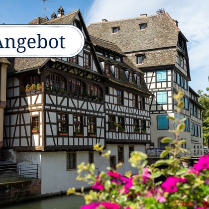 Traditionelle Fachwerkhäuser entlang eines Kanals mit rosa Blumen im Vordergrund. Textüberlagerung 'Angebot'.