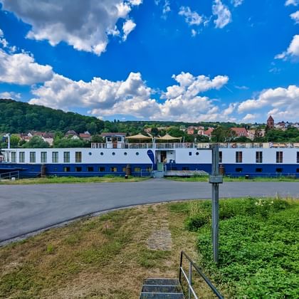 MS Casanova, ein weiß-blaues Flusskreuzfahrtschiff, am Flussufer angelegt mit historischer Stadt und bewaldeten Hügeln im Hintergrund.