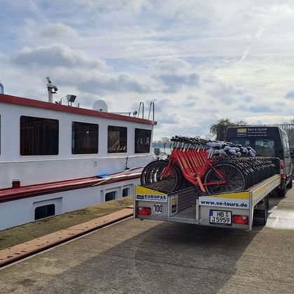 Lastwagen mit roten Leihfahrrädern neben einem weißen Flusskreuzfahrtschiff. Der Lkw zeigt se-tours.de Branding am Heck.