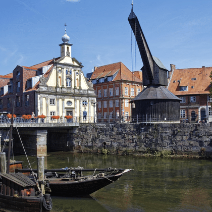 Alter hölzerner Hafenkran und Boot am Wasser in Lüneburg mit historischen Gebäuden, Kirchturm und Steinmauer entlang des Flusses.