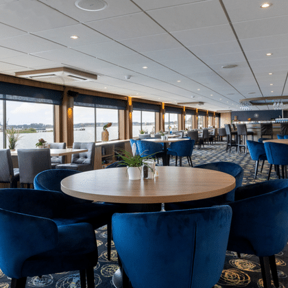 Modern ship lounge with blue velvet chairs around round tables, patterned carpet, and large windows overlooking a river. Bar area visible in background.