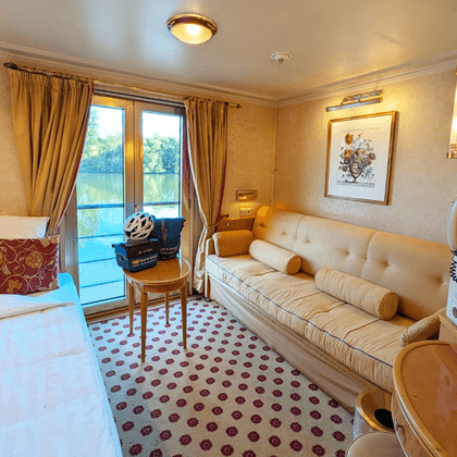 Bright cabin with white bed, beige sofa, patterned carpet, balcony door with river view, golden curtains, and mirror on Casanova upper deck.