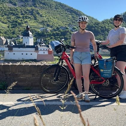 Two cyclists with red touring bikes posing on a road with Burg Pfalzgrafenstein castle on the Rhine River and green hills in the background.