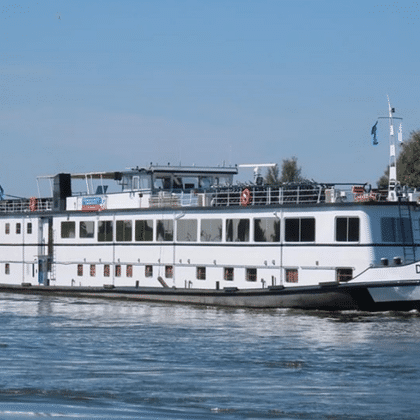 White river cruise ship De Nassau sailing on calm water. The multi-deck vessel has large windows and a sun deck, with green riverbanks visible.