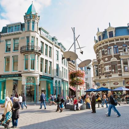 Pedestrian square in downtown Arnhem with historic buildings featuring turquoise corner building and ornate architecture. People walking across cobblestones.