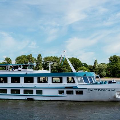 Weißes Flusskreuzfahrtschiff namens Switzerland fährt auf einem Fluss mit grünen Bäumen und blauem Himmel im Hintergrund. Das Schiff hat zwei Decks mit Fenstern.