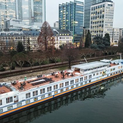 MS Classica river cruise ship with white hull and orange stripe moored at urban waterfront. Modern skyscrapers and bare trees visible in background.