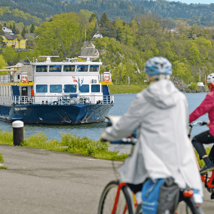 Zwei Radfahrer mit Helmen und Tourenrädern beobachten das Passagierschiff Prinzessin Katharina am Flussufer mit grünen Hügeln im Hintergrund.