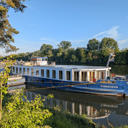 Blau-weißes Flusskreuzfahrtschiff Casanova an einem Holzsteg auf ruhigem Fluss, umgeben von grünen Bäumen unter blauem Himmel.