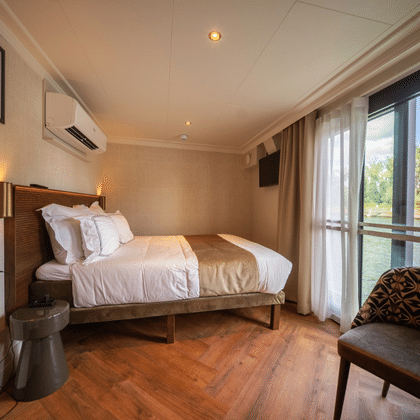 Moderne Suite auf der Magnifique X mit Doppelbett, Holzboden, Klimaanlage und großen Fenstern mit Blick auf einen Fluss mit grüner Landschaft.