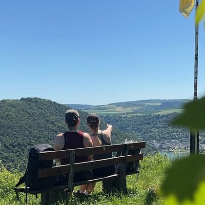 Zwei Wanderer sitzen auf einer Holzbank mit Blick auf Oberwesel und das Rheintal, umgeben von grünen Hügeln und Weinbergen unter blauem Himmel.