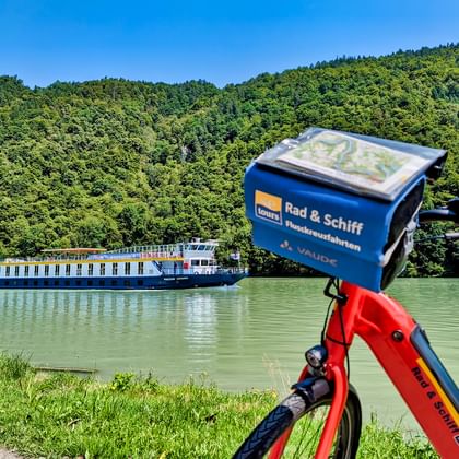 Blue and white river cruise ship on green water with forested hills. Red bicycle with blue SE-Tours bag in foreground.