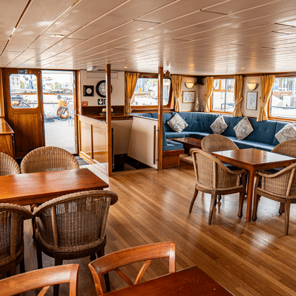 Heller Salon an Bord der Clair de Soleil mit Holzboden, Korbstühlen, blauem Sofa, goldenen Vorhängen und großen Fenstern mit Blick auf das Deck.