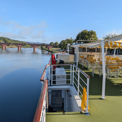 Sun deck of river cruise ship Casanova with yellow-white striped awning, wooden chairs, and green artificial turf. River and bridge visible in background.