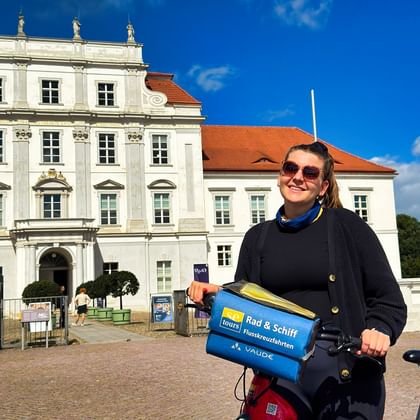Woman with bicycle and blue tour bag standing in front of the white baroque Oranienburg Palace under a bright blue sky.