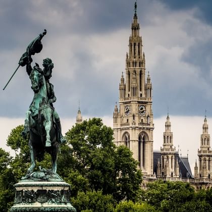 Bronzene Reiterstatue am Heldenplatz in Wien mit dem gotischen Turm und den Türmen des Wiener Rathauses im Hintergrund unter bewölktem Himmel.