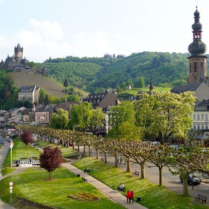 Panoramablick auf Cochem mit historischer Burg auf dem Hügel, barockem Kirchturm, Uferpromenade mit grünen Rasenflächen und bunten Häusern.