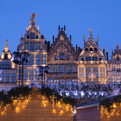 Historic guild houses at Antwerp's Grote Markt illuminated at dusk with Brabo fountain and Christmas market stalls decorated with lights.