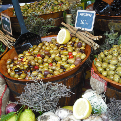 Holzfässer gefüllt mit verschiedenen marinierten Oliven auf einem Markt in der Provence. Preisschilder und frische Kräuter schmücken die Auslage.