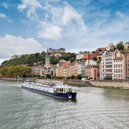Flusskreuzfahrtschiff Provence auf einem Fluss mit bunten historischen Gebäuden am Ufer und einer Hügelstadt unter blauem Himmel.