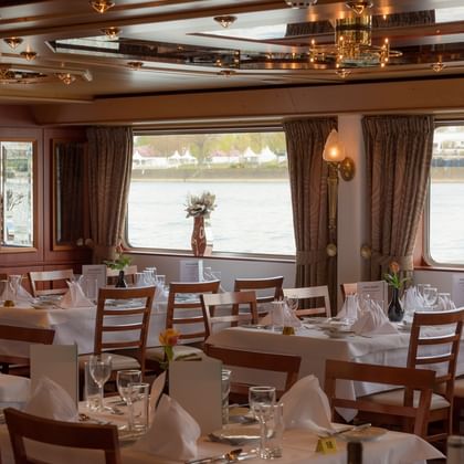 Elegantes Schiffsrestaurant mit weiß gedeckten Tischen, Holzstühlen und großen Fenstern mit Blick aufs Wasser. Braune Vorhänge und dekorative Decke.