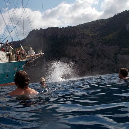 Menschen schwimmen im blauen Meerwasser nahe dem Segelschiff ATLANTIS mit grünem Rumpf. Felsige Küste im Hintergrund unter bewölktem Himmel.