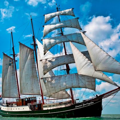 Traditionelles dreimastiges Segelschiff Simmer fan Fryslân mit voll gesetzten weißen Segeln auf ruhigem Wasser unter blauem Himmel mit Wolken.