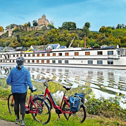 Radfahrer mit rotem Fahrrad beobachtet weißes Flusskreuzfahrtschiff. Burg auf Hügel mit Weinbergen im Hintergrund unter blauem Himmel.