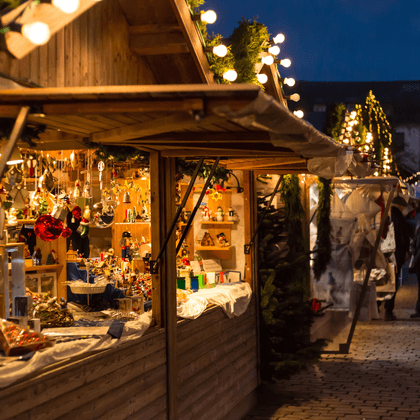 Hölzerner Weihnachtsmarktstand beleuchtet in der Dämmerung, mit festlicher Dekoration und Geschenken. Besucher gehen auf dem Kopfsteinpflaster.