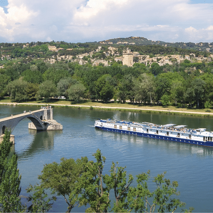 Blau-weißes Flusskreuzfahrtschiff Provence auf ruhigem Fluss an historischer Steinbrücke vorbei mit grünen Ufern und Hügelstadt im Hintergrund.