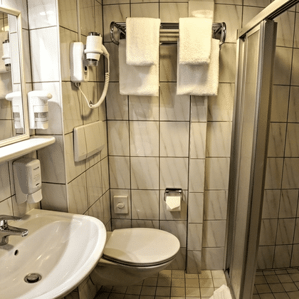 Kompaktes Badezimmer auf MS Classica mit beigen Fliesen, weißem Waschbecken, WC, Glasduschkabine, Wandhaartrockner und weißen Handtüchern.