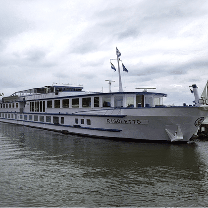 Weißes Flusskreuzfahrtschiff Rigoletto an einem Holzsteg unter bewölktem Himmel. Ein Segelboot ist im Hintergrund auf ruhigem Wasser sichtbar.