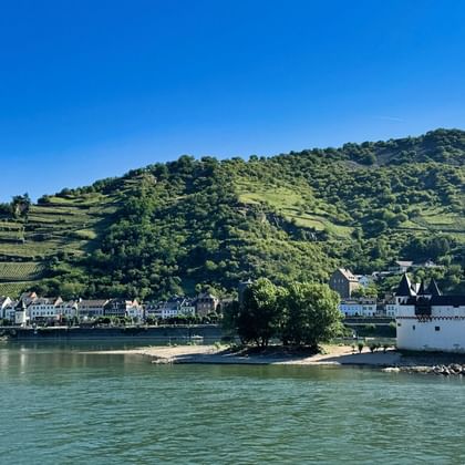 White fortress Pfalzgrafenstein on Rhine island with terraced vineyards on hillsides, a riverside town, and a castle ruin above.