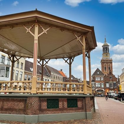 Wooden bandstand with ornate Victorian details on a brick base in Kampen's market square, surrounded by historic buildings and a church tower.