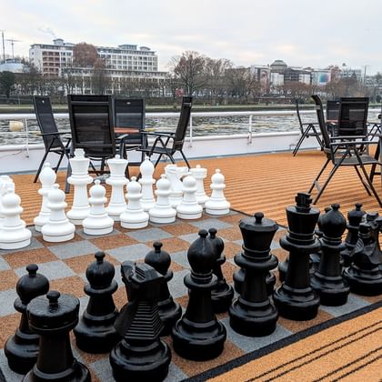 Sonnendeck der MS Classica mit übergroßem Schachspiel auf Holzdeck. Schwarze Klappstühle um das Brett mit Fluss und Stadtpanorama im Hintergrund.